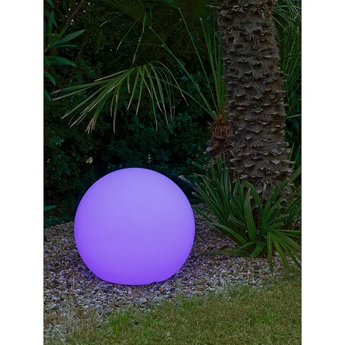 Starlux Bola solar Venus 30 - 0,5 W, 30 cm, RGB, 50 lm - Una bola de jardín LED púrpura brillante se asienta entre grava y vegetación, proporcionando iluminación ambiental para espacios exteriores.