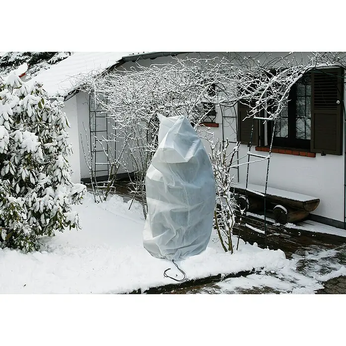Floraworld Haubenvlies - 2 Stk., 100 x 150 cm, Geeignet für: Überwinterung - Die Pflanze ist mit einer weißen Frostschutzhülle abgedeckt, um sie im Winter vor Kälte und Schnee zu schützen. Im Hintergrund befindet sich ein schneebedeckter Garten.