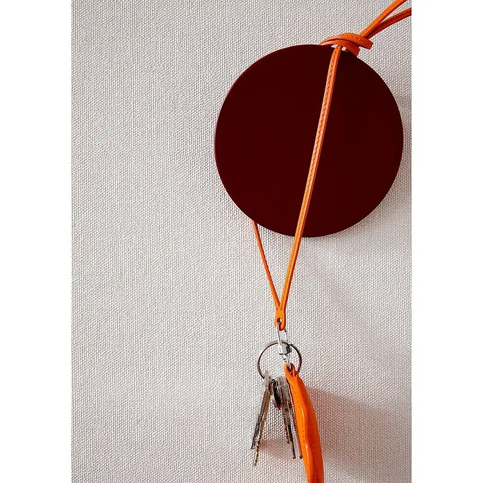 SCHÖNER WOHNEN-Kollektion Vliestapete Unistruktur - Greige, Uni, 10,05 x 0,53 m - Ein runder, weinroter Schlüsselhalter mit orangefarbenem Band und Schlüsselbund hängt an einer hellen, strukturierten Wand. Praktisch und stilvoll.