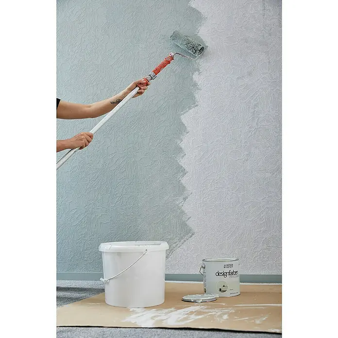 SCHÖNER WOHNEN-Kollektion Vliestapete - Weiß, Floral, 10,05 x 0,53 m - Ein Innenraumfoto zeigt einen Mann, der eine Wand mit einem Farbroller in einem sanften Blauton streicht, wobei ein Farbeimer und eine Dose mit der Aufschrift "Designfarben" auf dem Boden stehen.