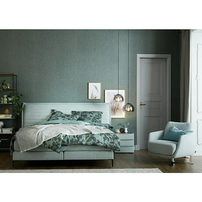 SCHÖNER WOHNEN-Kollektion Vliestapete Unistruktur - Grün/Grau, Uni, 10,05 x 0,53 m - Schlafzimmer mit einem grünen Bett mit Blumenmuster, einem Nachttisch und einer Leseecke mit einem grauen Sessel, der zum Entspannen einlädt.