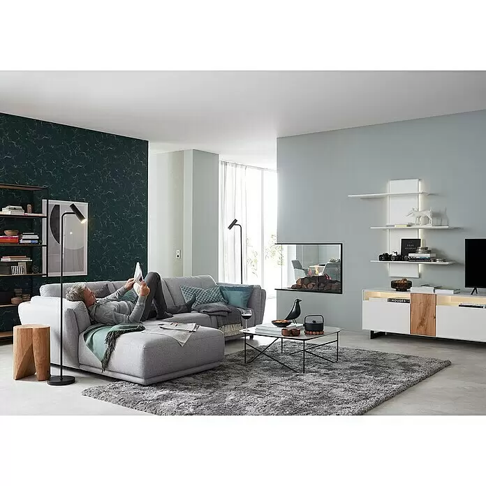 SCHÖNER WOHNEN-Kollektion Vliestapete Marmoroptik - Grün/Grau, Steinoptik, 10,05 x 0,53 m - Ein Mann entspannt auf einem grauen Stoff-Ecksofa mit Chaiselongue, liest Zeitung in einem modernen Wohnzimmer.