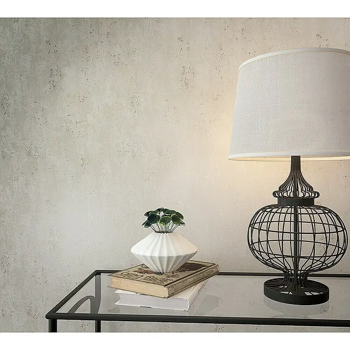 Marburg Sand & Stones Vliestapete City Glam I - Creme, Betonoptik, 10,05 x 0,53 m - Nahaufnahme einer Tischlampe mit einem Drahtfuß und einem weißen Lampenschirm, die auf einem Tisch mit einem Buch und einer Pflanze steht.