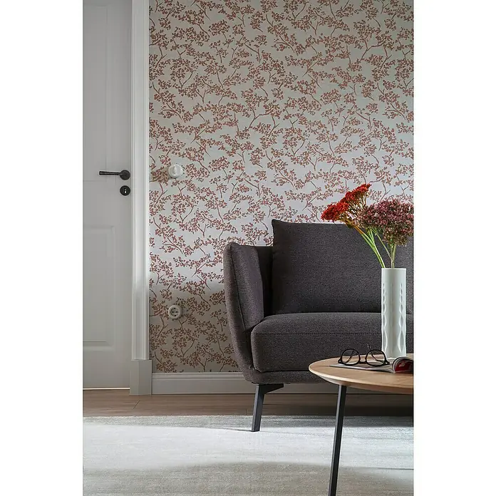 SCHÖNER WOHNEN-Kollektion New Spirit Vliestapete Blüten-Allover - Grau/Gold, Floral, 10,05 x 0,53 m - Nahaufnahme eines stilvollen grauen Sofas mit Blumenvase auf einem Holztisch, vor einer Wand mit floralem Muster.