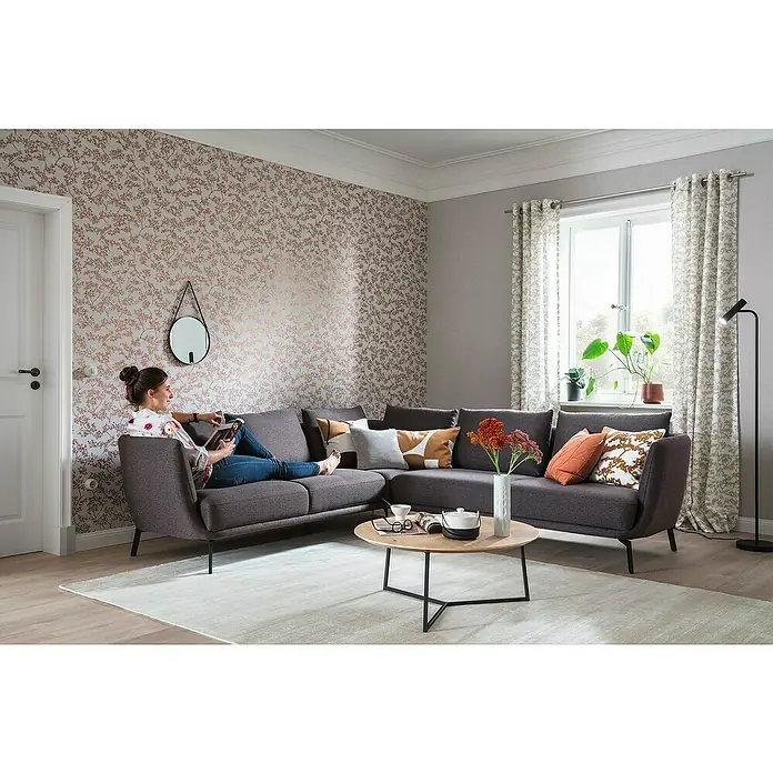 SCHÖNER WOHNEN-Kollektion New Spirit Vliestapete Blüten-Allover - Grau/Gold, Floral, 10,05 x 0,53 m - Einladendes Wohnzimmer mit einem grauen Ecksofa, das zum Entspannen einlädt. Der Teppich und die floralen Akzente schaffen eine gemütliche Atmosphäre.