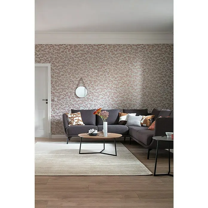 SCHÖNER WOHNEN-Kollektion New Spirit Vliestapete Blüten-Allover - Grau/Gold, Floral, 10,05 x 0,53 m - Helle Wohnung mit einer stilvollen grauen Eckcouch mit Zierkissen, einem runden Couchtisch und einem hellen Teppich auf Holzboden.