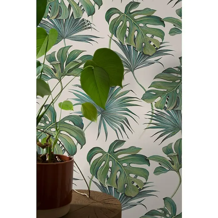 SCHÖNER WOHNEN-Kollektion New Spirit Vliestapete Fensterblatt - Grün/Weiß, Floral, 10,05 x 0,53 m - Zimmerpflanze in braunem Topf, abgebildet vor einer Wand mit tropischem Blättermuster. Die Pflanze ist grün und frisch.