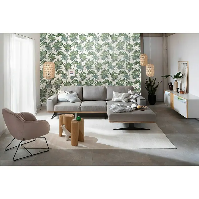 SCHÖNER WOHNEN-Kollektion New Spirit Vliestapete Fensterblatt - Grün/Weiß, Floral, 10,05 x 0,53 m - Das moderne Wohnzimmer verfügt über ein graues Ecksofa, Akzentmöbel aus Holz und eine dekorative grüne Blättertapete.