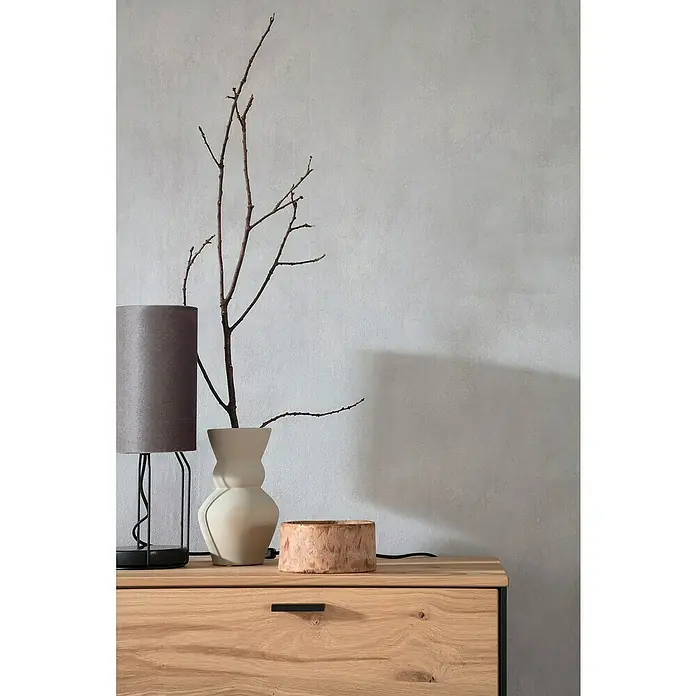 SCHÖNER WOHNEN-Kollektion New Spirit Vliestapete - Shadowgrau, Uni, 10,05 x 0,53 m - Dekorative Komposition mit Lampe, Vase mit Zweigen und Holzgefäß auf einem hellen Holzschrank vor einer grauen Wand.