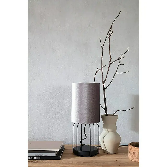 SCHÖNER WOHNEN-Kollektion New Spirit Vliestapete - Shadowgrau, Uni, 10,05 x 0,53 m - Nahaufnahme einer modernen Tischlampe mit grauem Lampenschirm und schwarzem Gestell auf einem Holztisch, neben einer Vase mit Zweigen.