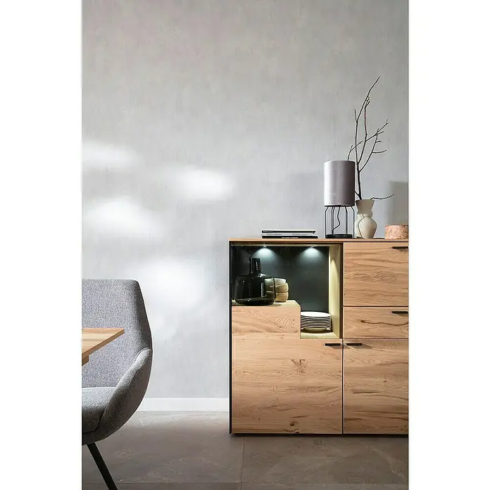 SCHÖNER WOHNEN-Kollektion New Spirit Vliestapete - Shadowgrau, Uni, 10,05 x 0,53 m - Ein stilvolles Sideboard aus hellem Holz mit Fächern und Schubladen, beleuchtet mit Dekorationsgegenständen. Perfekt für modernes Wohnen.