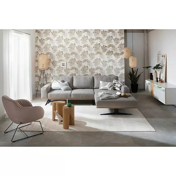 SCHÖNER WOHNEN-Kollektion New Spirit Vliestapete Fensterblatt - Beige/Weiß, Floral, 10,05 x 0,53 m - Ein modernes Wohnzimmer mit grauem Ecksofa, beigem Sessel und Holznebentischen auf hellem Teppich, ergänzt durch tropische Blatt-Tapete.