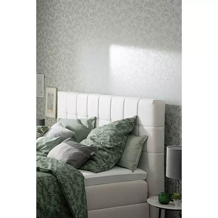 SCHÖNER WOHNEN-Kollektion New Spirit Vliestapete Blüten-Allover - Beige/Blau, Floral, 10,05 x 0,53 m - Ein Bett mit weißem gepolstertem Kopfteil und grün gemusterter Bettwäsche in einem Zimmer mit passender botanischer Tapete.