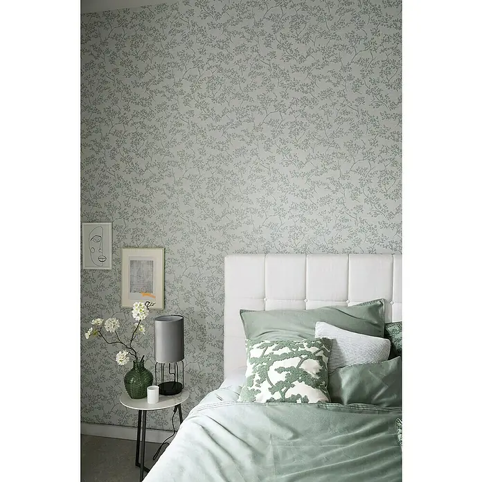 SCHÖNER WOHNEN-Kollektion New Spirit Vliestapete Blüten-Allover - Beige/Blau, Floral, 10,05 x 0,53 m - Ein Schlafzimmer mit einem mit grünem Blumendesign versehenen Bett, einem Nachttisch mit Lampe und Deko an der Wand.