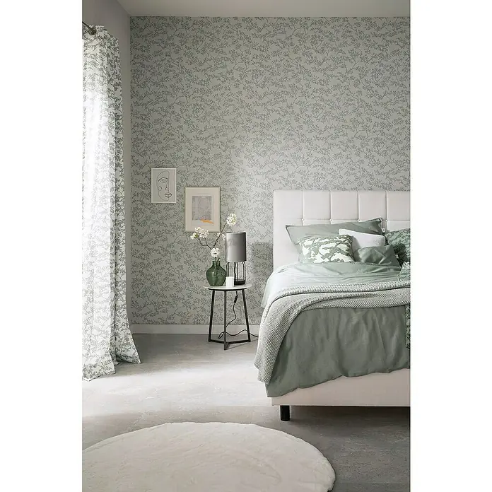 SCHÖNER WOHNEN-Kollektion New Spirit Vliestapete Blüten-Allover - Beige/Blau, Floral, 10,05 x 0,53 m - Ein Schlafzimmer mit einem Bett, Nachttisch und einer Lampe vor einer Wand mit Blumenmuster, hervorgehoben durch einen weichen, runden Teppich.