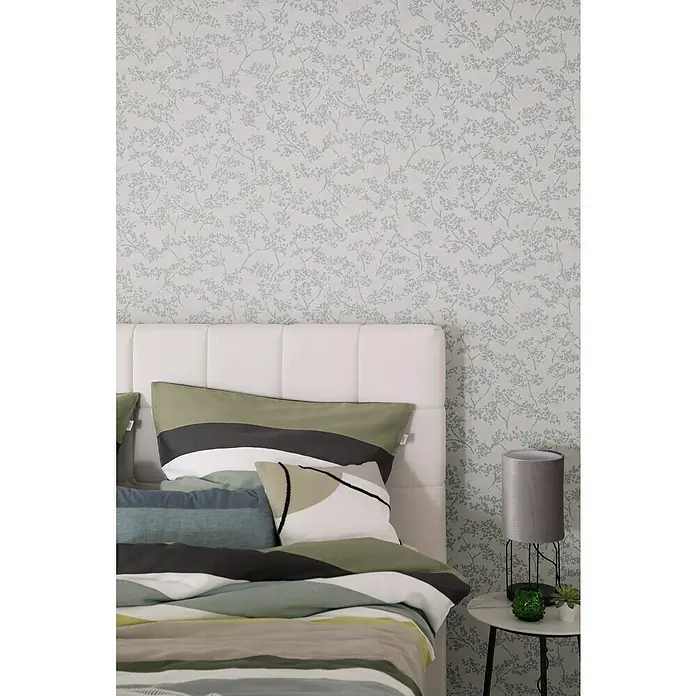 SCHÖNER WOHNEN-Kollektion New Spirit Vliestapete Blüten-Allover - Beige/Blau, Floral, 10,05 x 0,53 m - Nahaufnahme eines Schlafzimmers mit einem Bett, das mit Kissen und einer Tagesdecke in Grau-, Grün- und Weißtönen dekoriert ist.