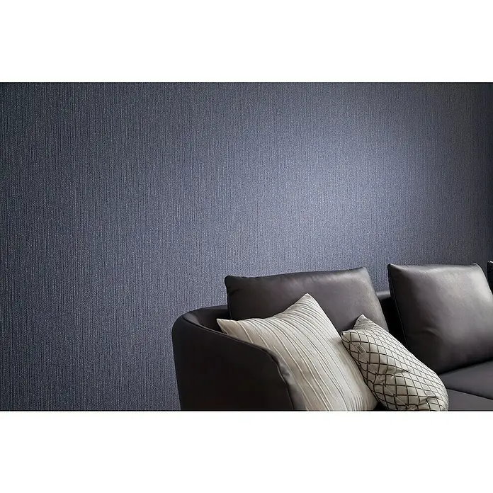 SCHÖNER WOHNEN-Kollektion New Spirit Vliestapete Struktur - Blau, Uni, 10,05 x 0,53 m - Nahaufnahme eines modernen grauen Sofas mit Kissen vor einer strukturierten blauen Wand. Das Sofa lädt zum Entspannen ein.