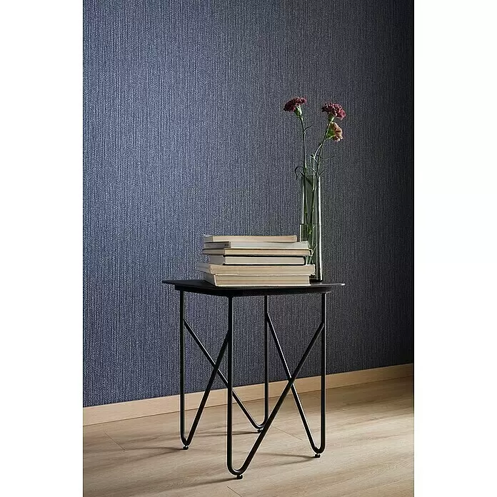 SCHÖNER WOHNEN-Kollektion New Spirit Vliestapete Struktur - Blau, Uni, 10,05 x 0,53 m - Ein schwarzer Beistelltisch aus Metall mit X-förmigen Beinen trägt Bücher und eine Vase mit roten Blumen vor einer strukturierten blauen Wand.