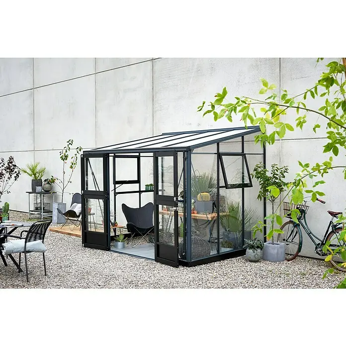 Juliana Gewächshaus Veranda - Außenmaß inkl. Dachüberstand (B x T): 221 x 296 cm, Material Seitenwand: Glas, 3 mm, Anthrazit - Das Bild zeigt ein elegantes Gewächshaus mit dunklem Rahmen, das in einem Garten mit Kiesboden und verschiedenen Pflanzen und Sitzgelegenheiten steht.