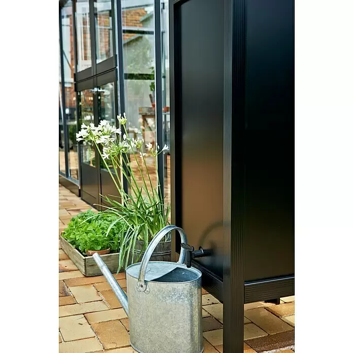 Juliana Regenspeicher - L x B x H: 46 x 46 x 127 cm, Schwarz - Schwarzer Regenwassertank mit Hahn, neben einer verzinkten Gießkanne und Pflanzen an einem Gewächshaus, zeigt seine Outdoor-Nutzung.