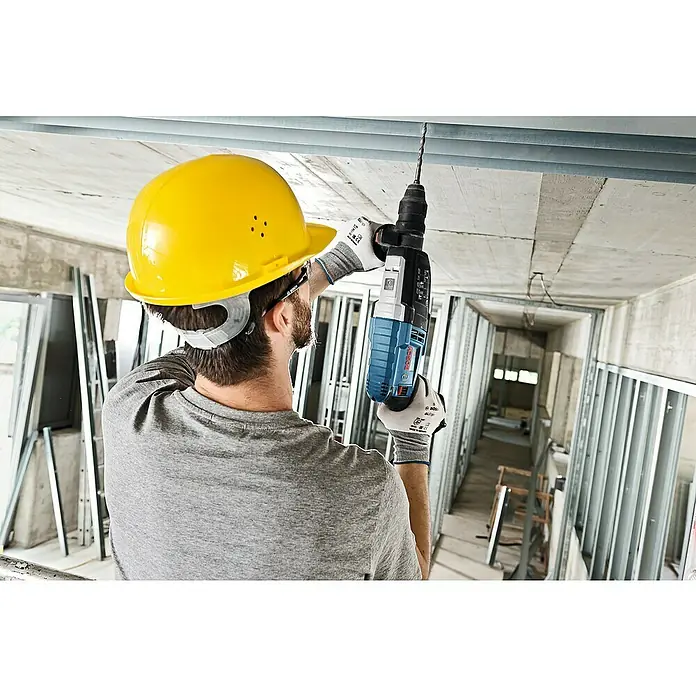 Bosch Professional Martillo combinado GBH 2-28 F - 880 W, 3,2 J, Alojamiento SDS-Plus, Incluye 3 brocas y cinceles - Un trabajador de la construcción con un casco amarillo perfora el techo con un taladro Bosch. El martillo perforador es azul y gris.