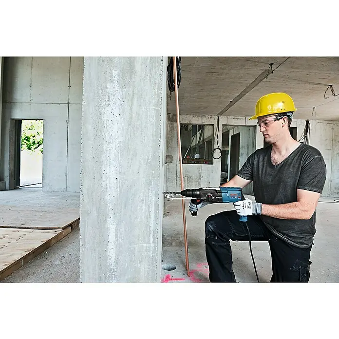 Bosch Professional Martillo combinado GBH 2-28 F - 880 W, 3,2 J, Alojamiento SDS-Plus, Incluye 3 brocas y cinceles - Un trabajador de la construcción se arrodilla y perfora una pared de hormigón con un martillo rotativo Bosch, con equipo de seguridad.
