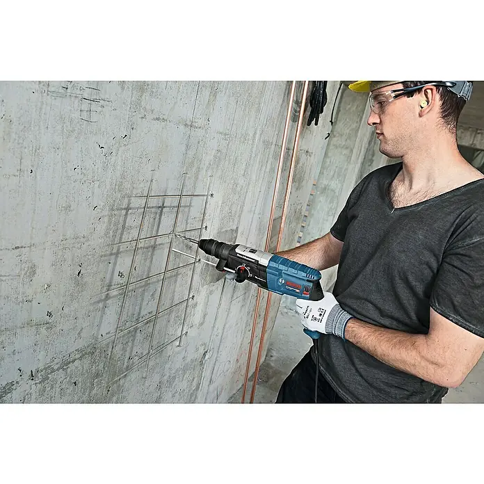 Bosch Professional Martillo combinado GBH 2-28 F - 880 W, 3,2 J, Alojamiento SDS-Plus, Incluye 3 brocas y cinceles - Un trabajador de la construcción usa un martillo perforador Bosch para perforar un agujero en una pared de hormigón con barras de refuerzo.