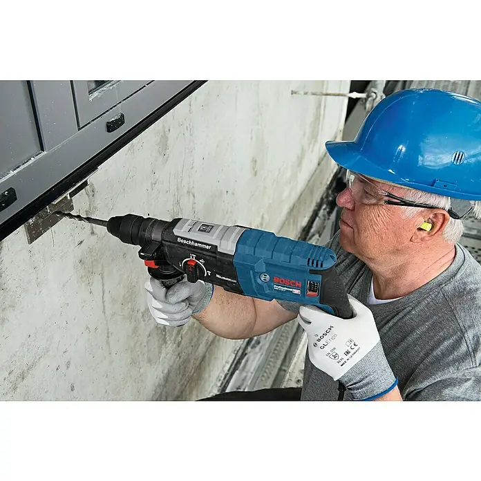 Bosch Professional Martillo combinado GBH 2-28 F - 880 W, 3,2 J, Alojamiento SDS-Plus, Incluye 3 brocas y cinceles - Un obrero de la construcción perfora una pared de hormigón con un Boschhammer Professional, protegido por casco y guantes.