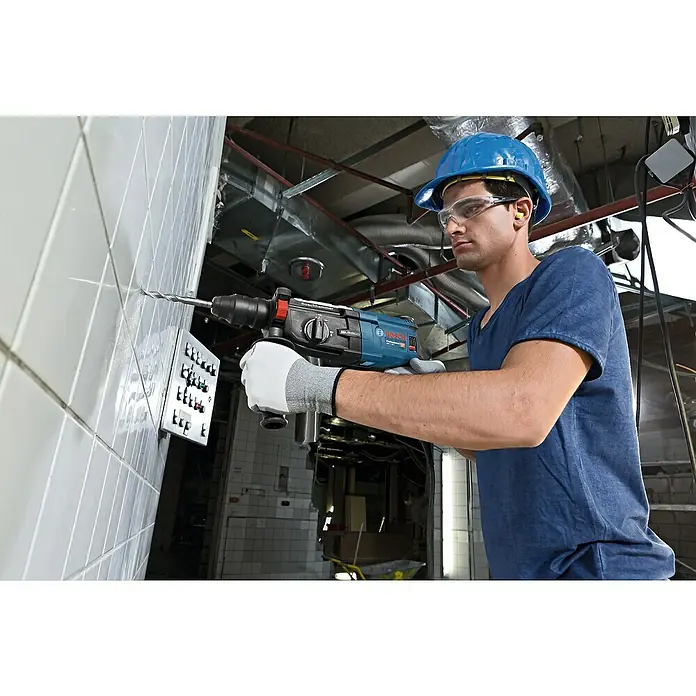 Bosch Professional Martillo combinado GBH 2-28 F - 880 W, 3,2 J, Alojamiento SDS-Plus, Incluye 3 brocas y cinceles - Un trabajador de la construcción con casco y gafas de seguridad perfora una pared con un martillo perforador Bosch.