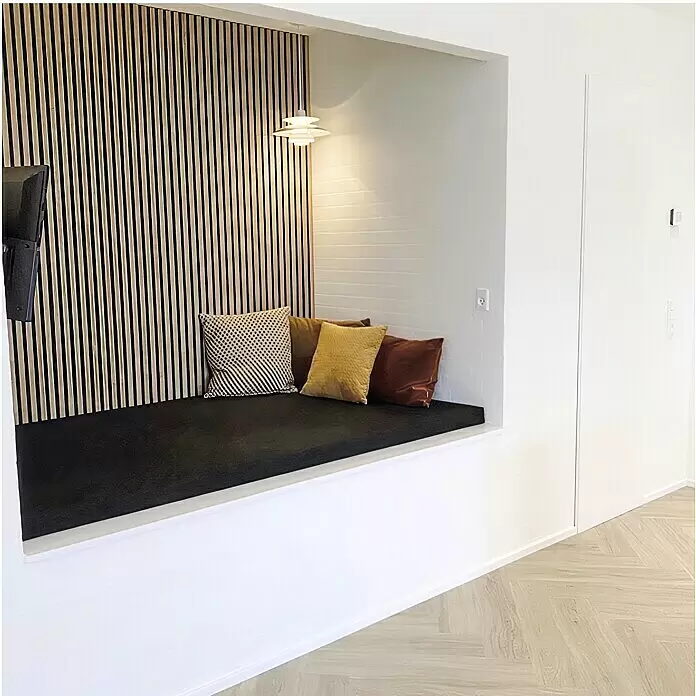FibroTech Akustikpaneel Premium - Light Oak, 2.440 x 605 x 22 mm - Eingebaute Sitznische mit schwarzem Kissen und drei Zierkissen vor einer Holzlamellen-Rückwand, beleuchtet von einer Pendelleuchte.
