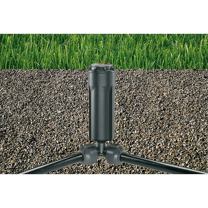 Gardena Sprinklersystem Winkelstück - Durchmesser: 25 mm, Größe Anschluss: ½″ - Nahaufnahme eines versenkregners, der zwischen Gras und Kies installiert ist. Das Bewässerungssystem ist einwandfrei und betriebsbereit.