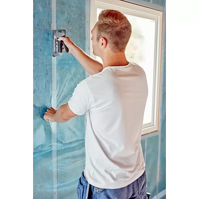 Rapid Grapadora manual ALU840 - Profundidad de grapado: 6 mm - 14 mm, Apto para: Grapas de alambre plano número 140 - Un hombre usa una grapadora gris con mango negro para fijar láminas protectoras azules a un marco de pared, mostrando su uso en aislamiento.
