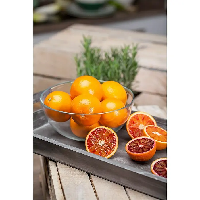 Piardino Árbol frutal Naranjo - Citrus x sinensis, Volumen de la maceta: 10 l - Un tazón de naranjas sanguinas de color naranja brillante se asienta sobre una bandeja de madera rústica, con frutas cortadas que muestran sus interiores rojos.
