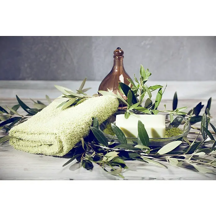 Piardino Olivenbaum - Olea europaea Florida, Topfgröße: 35 cm - Nahaufnahme eines hellgrünen Handtuchs, einer Seife in einer Schale, einer braunen Flasche und Olivenzweigen für ein Wellness-Erlebnis.