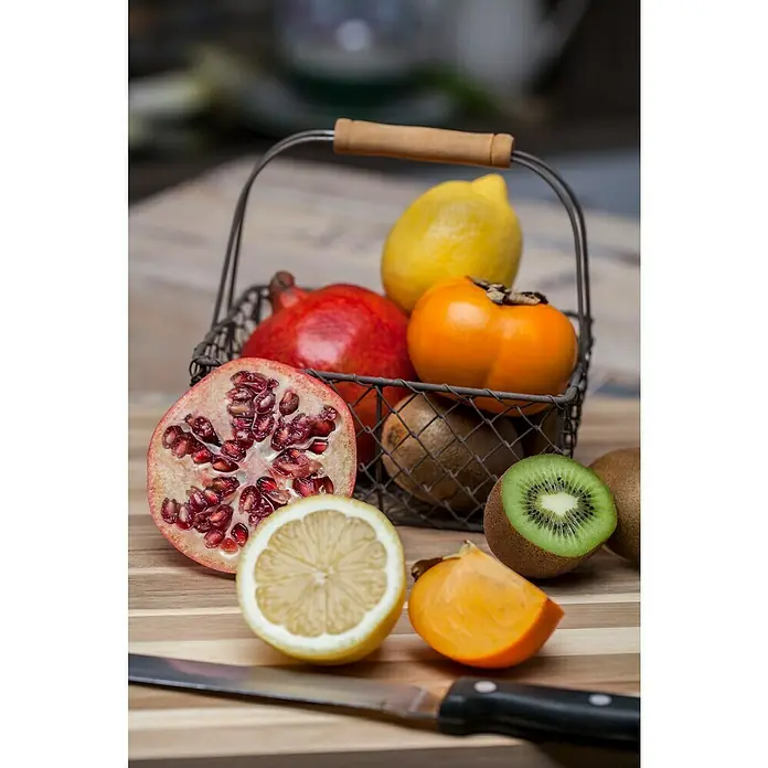 Granado - Punica granatum - Una cesta llena de frutas frescas como granada, limón, kiwi y caqui, parcialmente cortadas, sobre una mesa de madera con un cuchillo.