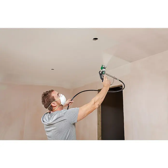 Wagner High Efficiency Airless Farbsprühsystem Control Pro 250 M - 550 W, Förderleistung: 1,25 l/min - Ein Mann mit Maske sprüht Farbe mit einer Farbspritzpistole an die Decke eines Raumes. Die Spritzpistole ist gut sichtbar.