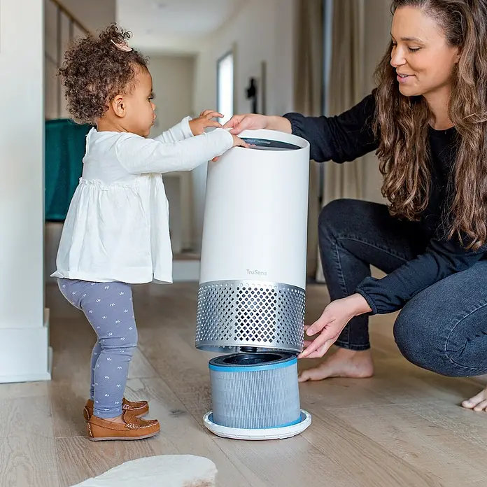 Leitz TruSens Aktivkohlefilter AllergiePassend für: TruSens Luftreiniger Z-2000/Z-2500, Höhe: 14,2 cm - Eine Frau und ein Kleinkind inspizieren einen TruSens Luftreiniger, der auf einem Holzboden steht, wobei der Fokus auf dem innovativen Filterdesign liegt.