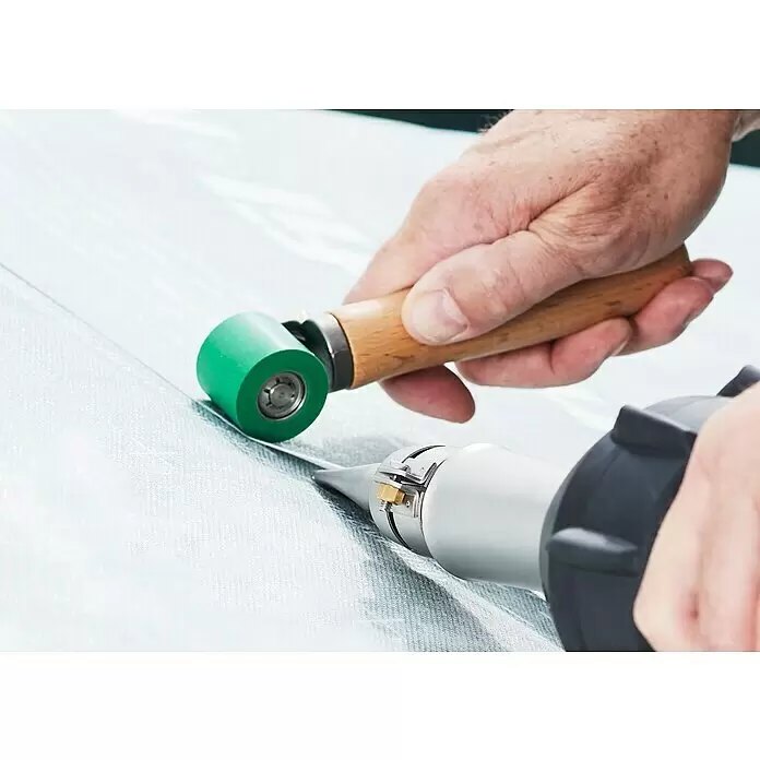 Leister Andruckrolle - Breite: 40 mm - Nahaufnahme einer Person, die eine Heißluftpistole und eine Rolle mit grünem Rad zum Schweißen oder Versiegeln eines hellen Materials verwendet.