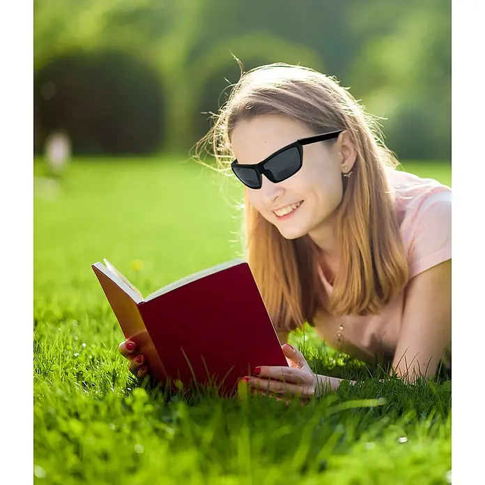 Magic Vision LED-Sonnenlesebrille - Schwarz, Vergrößerung 1: 300 % - Frau liegt im Gras und liest ein Buch mit einer schwarzen Sonnenbrille. Die Sonnenbrille bietet UV-Schutz und Komfort beim Lesen im Freien.