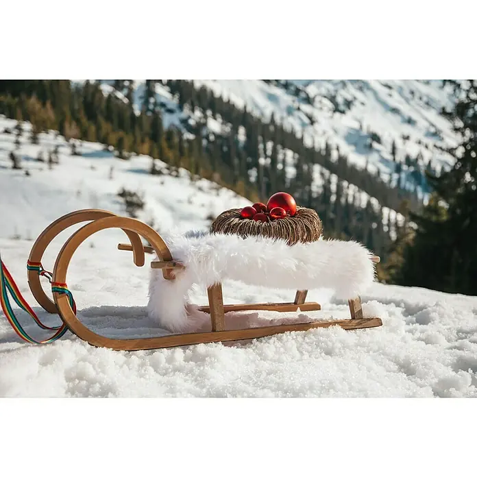 Esbeco Schaffell - Weiß, 80 x 50 cm, 100 % Echtfell - Festlicher Holzschlitten im Schnee, dekoriert mit weißen Fellen und roten Weihnachtskugeln, vor einer verschneiten Bergkulisse.