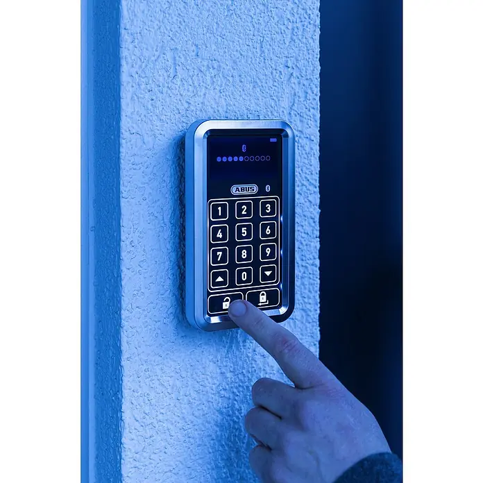 Abus HomeTec Pro Funk-Tastatur CFT 3100WL x B x H: 29 x 78 x 142 mm, Passend für: Abus HomeTec Pro Funk-Türschlossantrieb CFA3100, Weiß - Nahaufnahme eines ABUS-Türschloss-Keypads mit Fingerabdrucksensor und beleuchteten Tasten, das an einer Wand montiert ist.