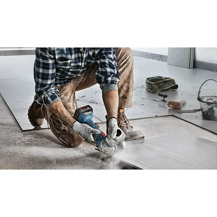 Bosch Professional AMPShare 18V Amoladora angular de batería GWS 18V-7 - 2 baterías, 4 Ah, Diámetro disco: 125 mm - Un trabajador de la construcción está arrodillado cortando baldosas con una amoladora angular Bosch, con polvo volando. Lleva ropa protectora.