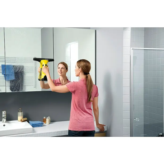 Kärcher Limpiadora de cristales de batería WV2 Plus N - Sin batería, Volumen de recipiente: 0,1 l - Mujer limpiando un espejo con un limpiacristales Kärcher, que asegura una limpieza sin rayas. El dispositivo es amarillo y negro.