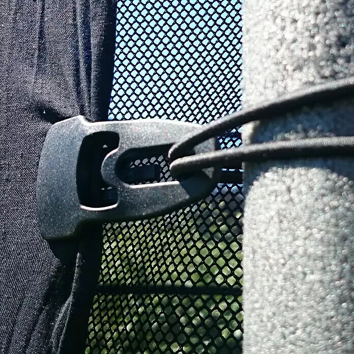 Clips Holdon Midi - 4 Stk., Schwarz - Schwarze Kunststoffschnalle mit Gummikordel, die schwarzen Stoff an einem strukturierten Pfosten neben einem Netzgewebe befestigt.