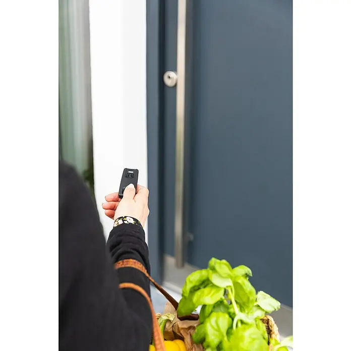 Abus HomeTec Pro Funkfernbedienung CFF3100 - Passend für: Abus HomeTec Pro Funk-Türschlossantrieb CFA3100 - Die Person öffnet mit einer Fernbedienung die Tür, während sie einen Korb mit Grünzeug hält. Moderne Haustechnik für Komfort und Sicherheit.