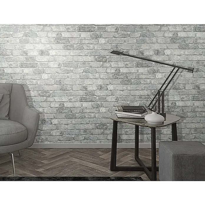 Marburg Sand & Stones Vliestapete Steinmauer II - Grauweiß, Steinoptik, 10,05 x 0,53 m - Elegante Inneneinrichtung mit grauem Sessel, Tisch mit Lampe und Büchern vor einer Backsteinmauer. Das stilvolle Design verleiht dem Raum ein modernes Ambiente.