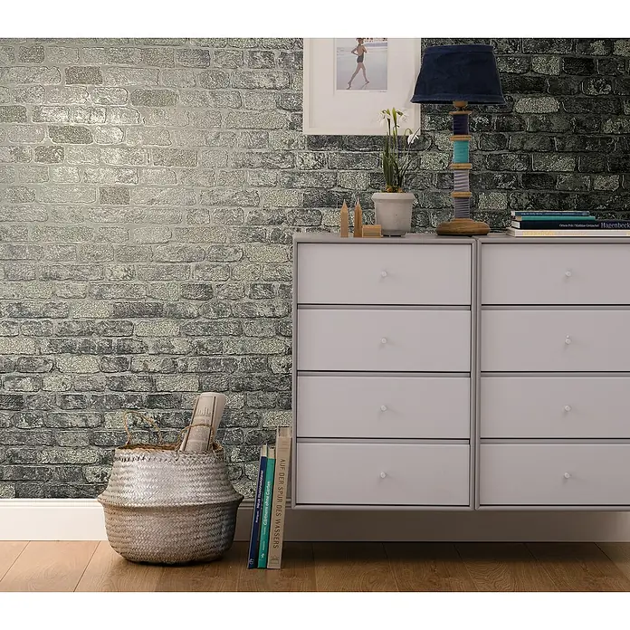 Marburg Sand & Stones Vliestapete Steinmauer I - Grau/Dunkelgrau, Steinoptik, 10,05 x 0,53 m - Eine moderne Kommode mit sechs Schubladen, platziert vor einer Backsteinmauer mit dekorativer Lampe und Büchern.