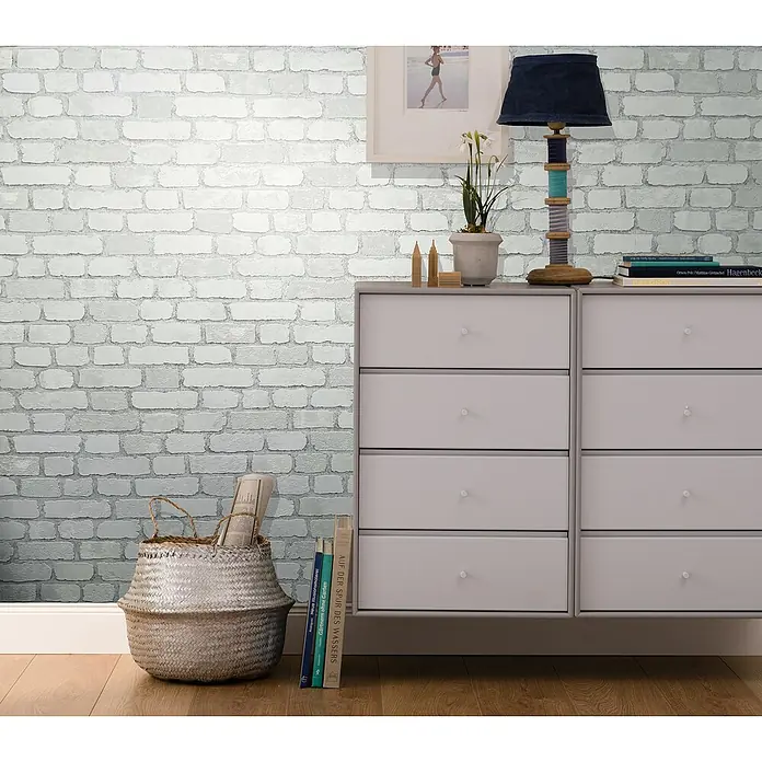 Marburg Sand & Stones Vliestapete Steinmauer II - Weiß, Steinoptik, 10,05 x 0,53 m - Kommode mit sechs Schubladen und einer Lampe oben drauf, die auf einem Holzboden steht und vor einer Ziegelwand platziert ist.