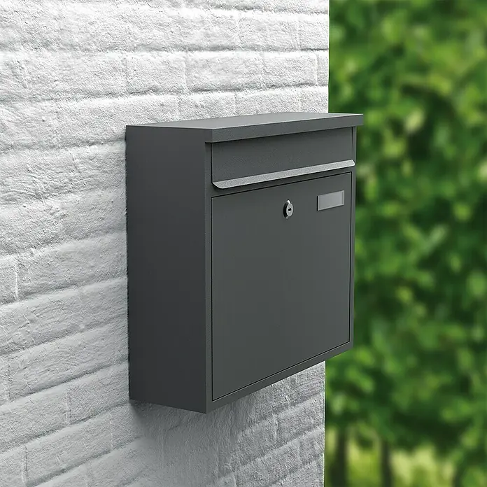 Basi Wandbriefkasten BK 360 M - L x B x H: 11,6 x 36 x 31,5 cm, Anthrazit, Stahl, Namensschild - Grauer Briefkasten an einer weißen Ziegelwand mit Bäumen im Hintergrund. Der Briefkasten ist schlicht und modern gestaltet.