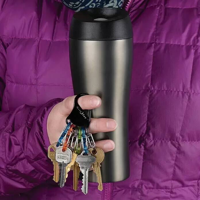 Nite Ize Mosquetón KeyRack - L x An: 12 x 2 mm, Acero inoxidable - Una persona sostiene una taza de viaje de acero inoxidable cepillado con tapa negra y un llavero mosquetón Nite Ize negro con varias llaves, vistiendo una chaqueta morada.